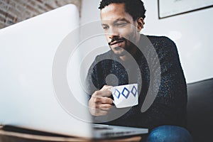 Cheerful African man using computer and smiling while sitting on the sofa.Black guy holding ceramic cup in hand.Concept