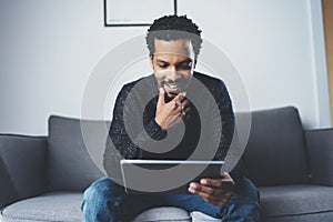 Cheerful African man using pc tablet and smiling while sitting on the sofa in his modern room.Concept of young business