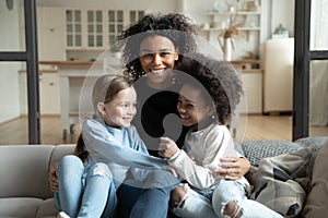 Cheerful African mother sitting on couch embraces multiracial daughters