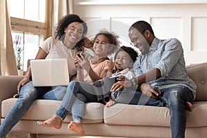 Cheerful african parents and kids laugh using devices on sofa photo