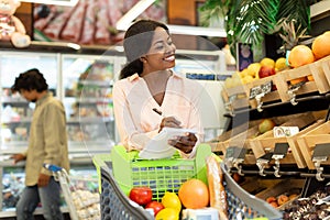 Cheerful African Woman Doing Grocery Shopping With List In Supermarket