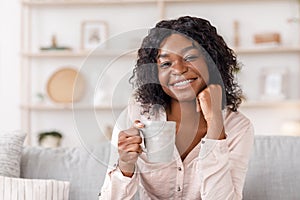 Cheerful african woman relaxing on couch at home with cup of coffee