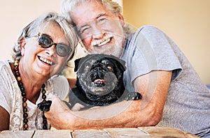 Cheerful alternative family with old senior couple people and funny black pug dog hug together in love activity - portrait of