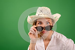 Cheerful amazed woman in a hat and sunglasses in a white shirt on a green background. Looks into the camera with his glasses down