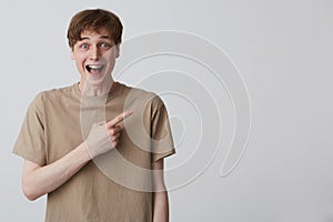 Cheerful amazed young man student with metal braces on teeth wears beige t shirt and points to the side  over white