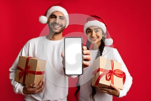 Cheerful Arab Couple Holding Christmas Presents And Showing Blank Smartphone