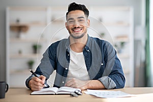 Cheerful arab guy in casual sitting at workdesk, taking notes