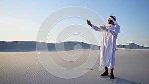 Cheerful Arabian Sheikh male tourist calls friend on videocall by cell and shows surroundings of white sand desert on
