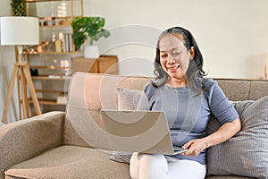 Cheerful Asian-aged retired woman enjoys watching movie on her laptop in her living room