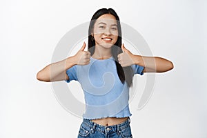 Cheerful asian brunette shows thumbs up and smiles, approves smth good, praising your choice, standing in tshirt over