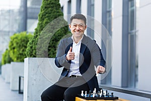 Cheerful Asian businessman playing chess in the yard smiling and looking forward to the camera