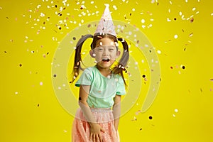 Cheerful Asian Child Girl Celebrating Birthday with Confetti on Yellow Isolated Background