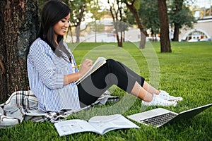 Cheerful asian female student in striped shirt, writing in notebook, while studying in park, outdoor