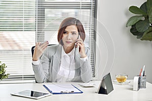 Cheerful asian female talking on mobile phone while sitting on desk with laptop. Business woman in casuals making a phone call and photo