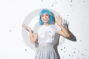 Cheerful asian girl in blue wig throwing confetti over white background, celebrating halloween, waving hand to say hello