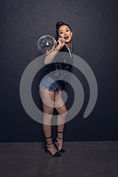 Cheerful asian girl holding disco ball and posing at grey wall in studio