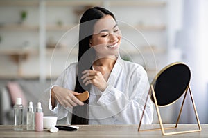 Cheerful asian lady combing hair with wooden brush