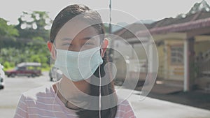 Cheerful Asian long hair pretty girl in protective mask wearing earphones standing on the street in residential area.