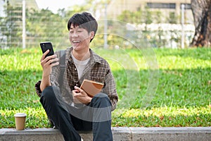 A cheerful Asian male college student sits on the street using his phone, laughing at messages