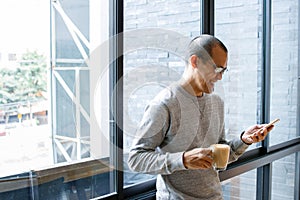 Cheerful asian male small business owner with coffee cup in hands searching on phone, reading incoming sms message on smartphone w