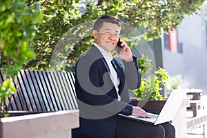 Cheerful asian man businessman working on laptop and talking on the phone, smiling sitting on a park bench near the office on