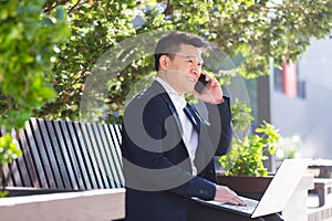 Cheerful asian man businessman working on laptop and talking on the phone, smiling sitting on a park bench near the office on