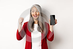 Cheerful asian mature woman showing blank smartphone screen and credit card, concept of e-commerce