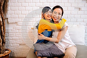 Cheerful Asian mother and daughter embracing while sitting on sofa