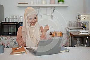 A cheerful Asian Muslim woman is looking at her laptop screen with a happy face