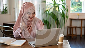 A cheerful Asian-Muslim woman is looking at her laptop screen with a happy face, good news