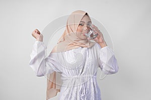 Cheerful Asian Muslim woman wearing white dress and hijab drinking a glass of water with a happy successful expression and gesture