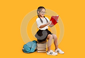 Cheerful Asian Schoolgirl Reading Sitting On Book Stack In Studio