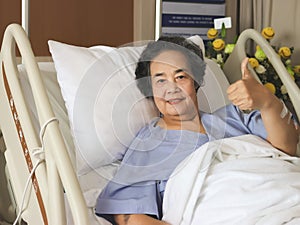 cheerful Asian senior female patient lying in hospital bed, smiling and showing her thumb up. Elderly health concept