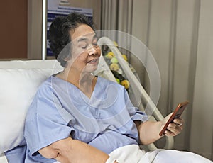 cheerful Asian senior female patient sitting in hospital bed, smiling and looking at mobile phone, video calling to her