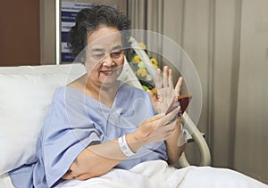 Cheerful  Asian senior  female patient  sitting  in hospital bed,  smiling and looking at mobile phone, video calling  and waving