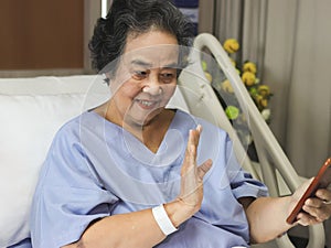 cheerful Asian senior female patient sitting in hospital bed, smiling and looking at mobile phone, video calling and waving