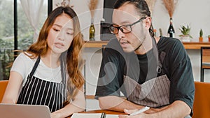 Cheerful Asian small business owners couple using laptop discussing project and finance with documents of shop for the month at