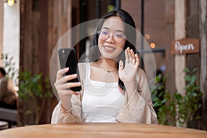 A cheerful Asian woman enjoys talking on a video call while sitting at an outdoor table of a cafe