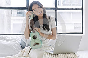 Cheerful asian woman with ukulele, laptop, pillows sitting on white bed beside window at apartment. Joyful young girl holding