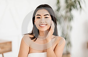 Cheerful asian woman wrapped in towel touching her face