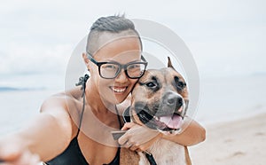 Cheerful asian young woman in eyeglases sitting and doing sekfie with her dog on the beach