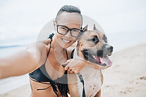 Cheerful asian young woman in eyeglases sitting and doing sekfie with her dog on the beach
