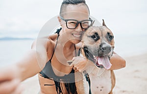 Cheerful asian young woman in eyeglases sitting and doing sekfie with her dog on the beach