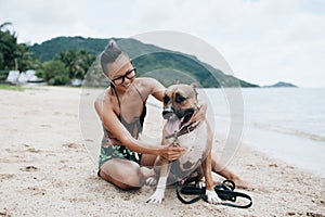 Cheerful asian young woman in eyeglases sitting and hugging her dog on the beach