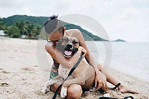 Cheerful asian young woman in eyeglases sitting and hugging her dog on the beach