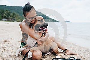 Cheerful asian young woman in eyeglases sitting and hugging her dog on the beach