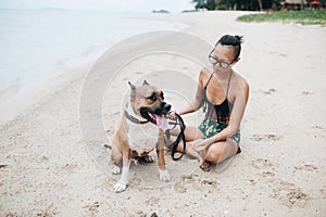 Cheerful asian young woman in eyeglases sitting and hugging her dog on the beach