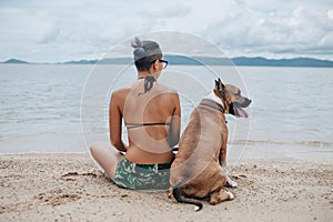 Cheerful asian young woman in eyeglases sitting and hugging her dog on the beach