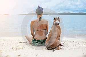 Cheerful asian young woman in eyeglases sitting and hugging her dog on the beach