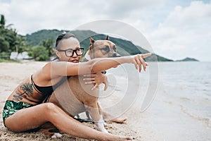 Cheerful asian young woman in eyeglases sitting and hugging her dog on the beach